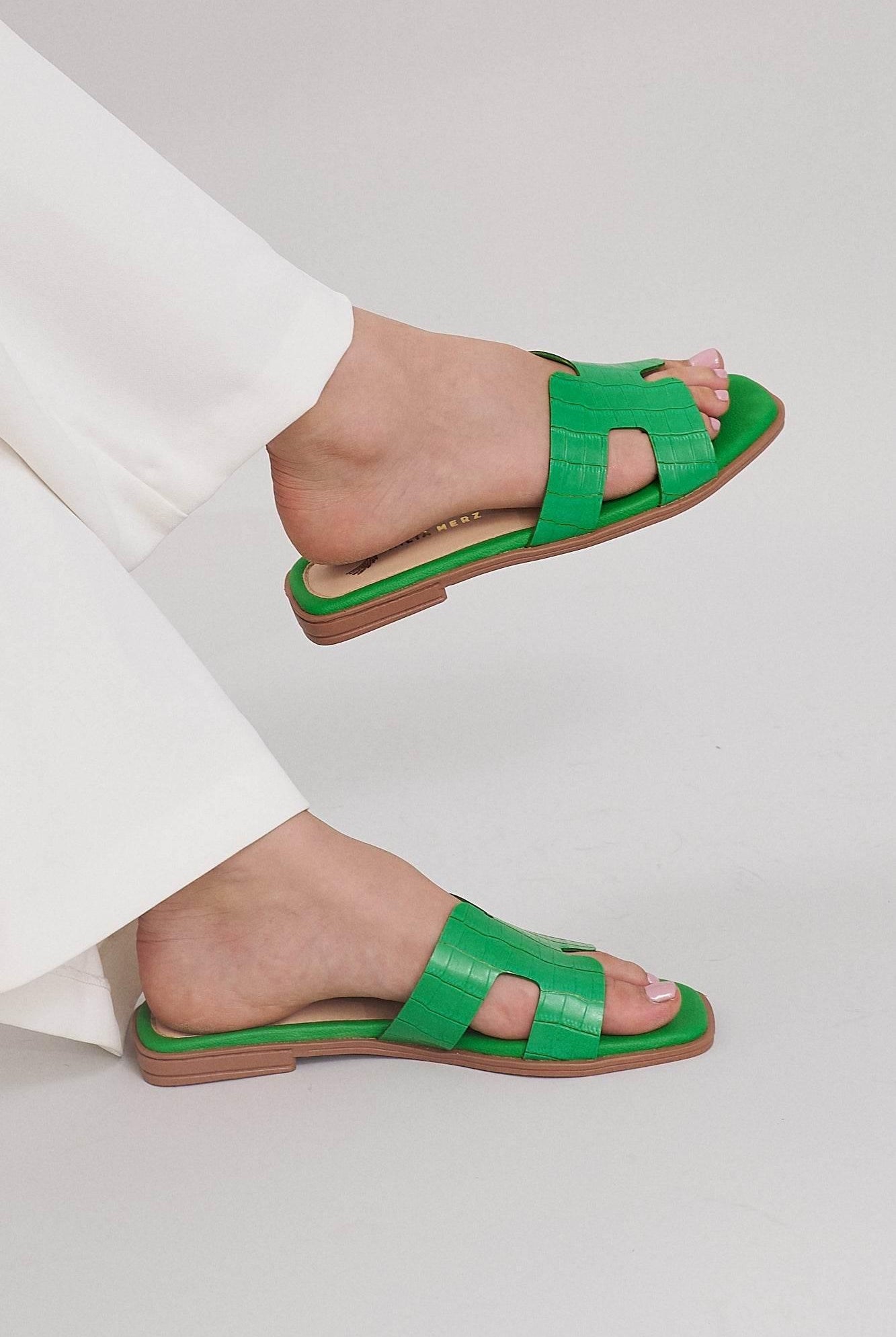 Green croc-embossed leather slide sandals on feet, paired with white pants