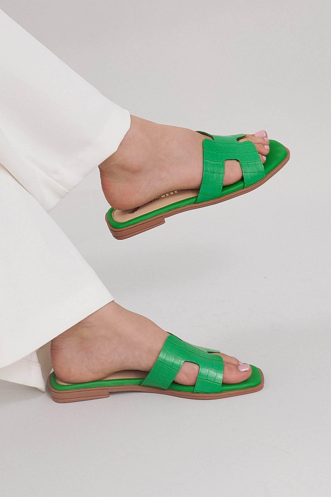 Green croc-embossed leather slide sandals on feet, paired with white pants