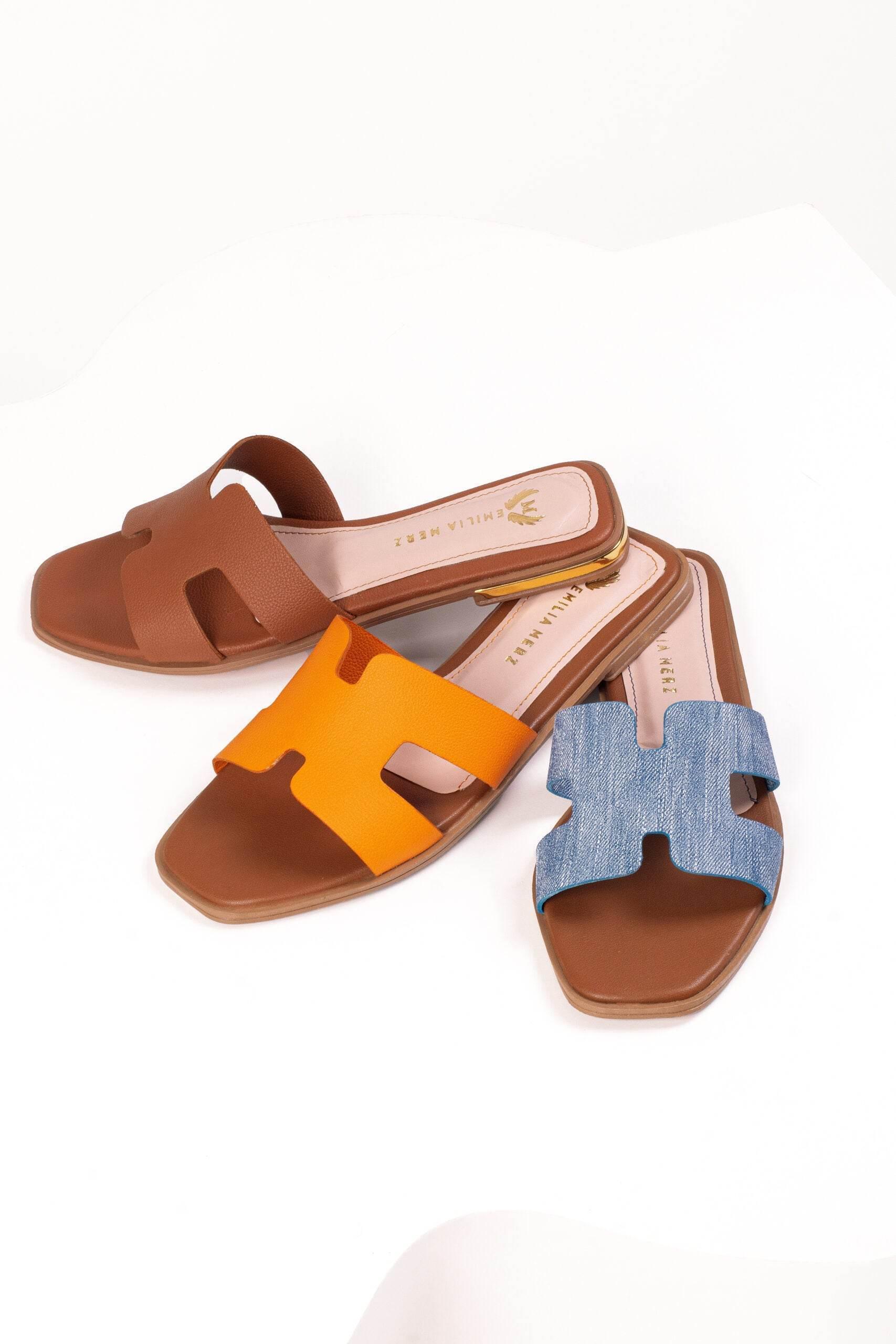 Three pairs of flat slide sandals in brown, orange, and denim blue on white background