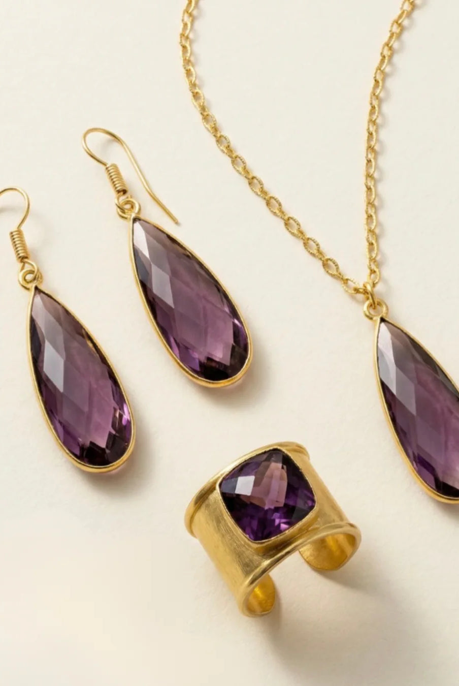Emilia Merz Aurea jewelry set featuring a pair of faceted deep violet drop earrings, a matching pendant necklace, and a cuffed statement ring set in 18K gold plating on a light background.