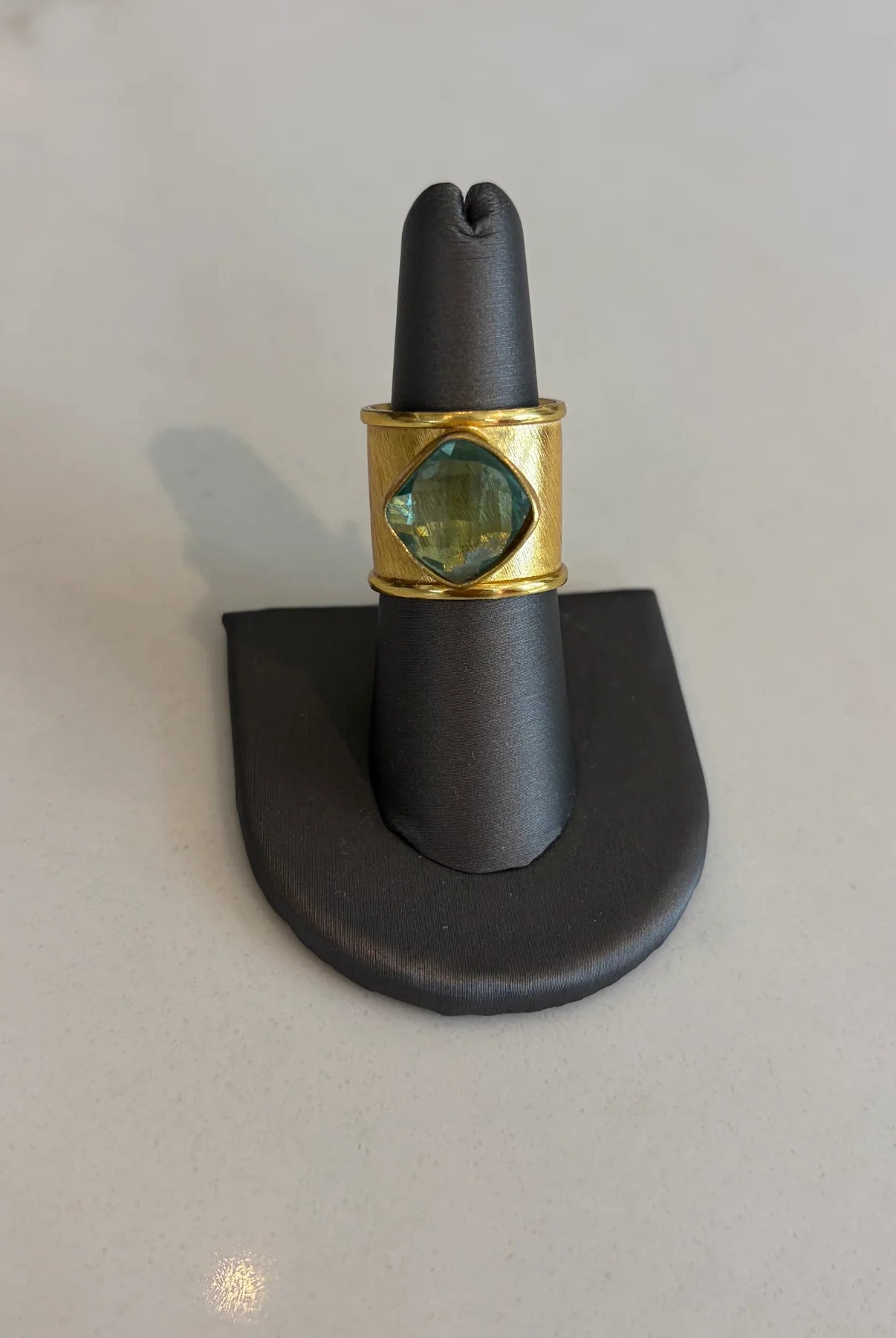 Gold aurea ring with a green gemstone on a black stand against a light gray background