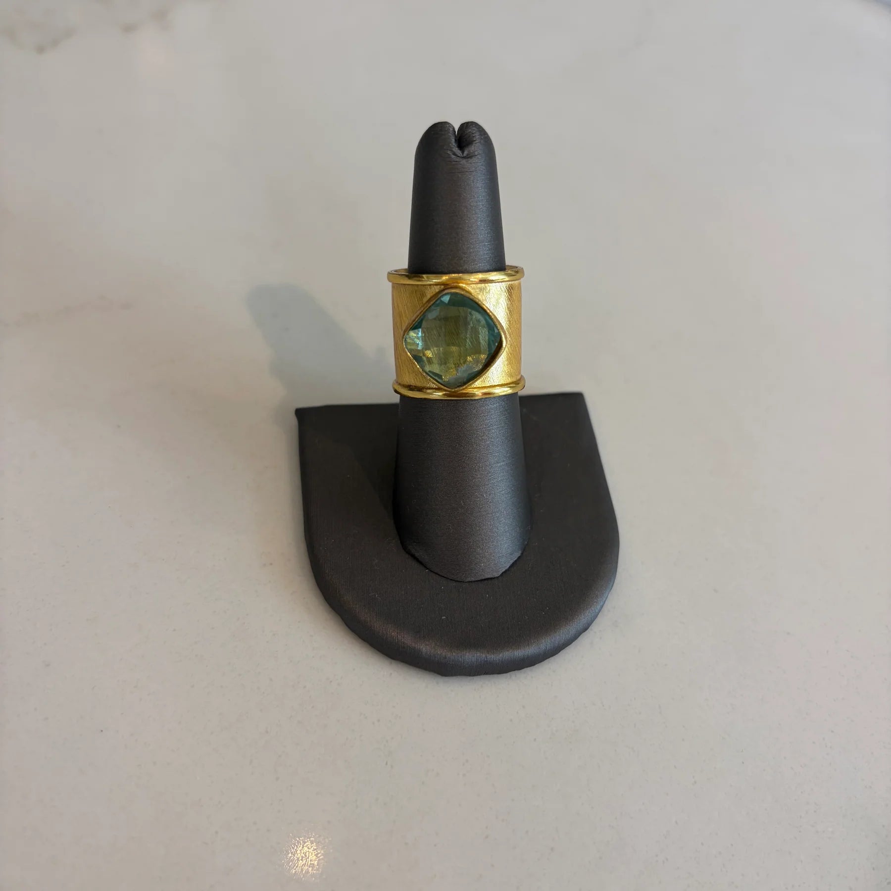 Gold aurea ring with a green gemstone on a black stand against a light gray background