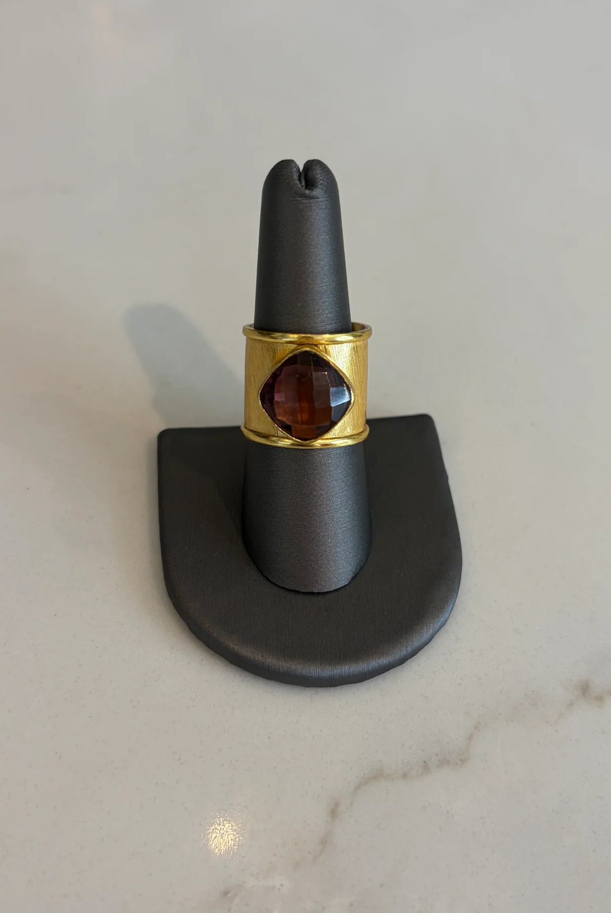 Front view of the Emilia Merz Aurea ring featuring a deep violet cushion-cut faceted stone set in a wide, cuffed 18K gold-plated band on a gray jewelry display.