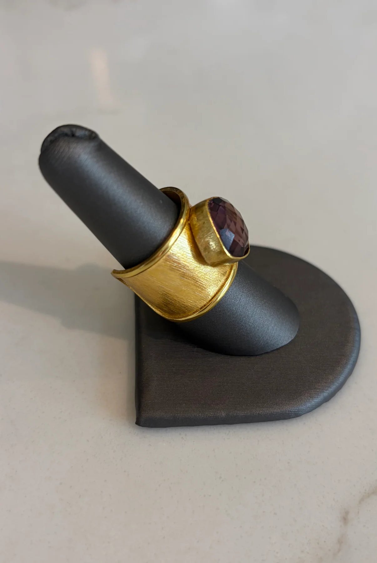 Side profile of the Emilia Merz Aurea statement ring showcasing the textured, adjustable gold band and deep violet faceted stone, displayed on a ring stand.