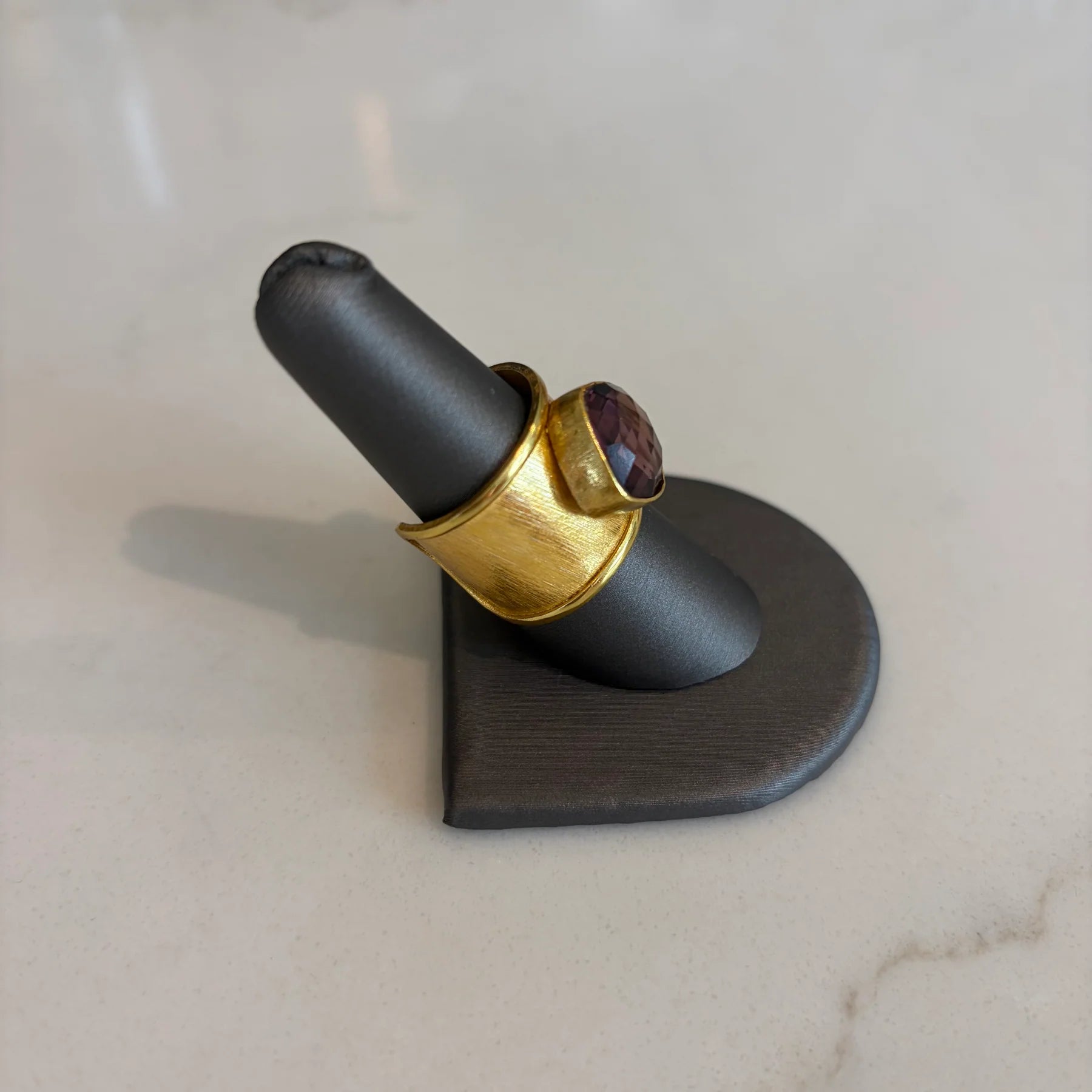 Side profile of the Emilia Merz Aurea statement ring showcasing the textured, adjustable gold band and deep violet faceted stone, displayed on a ring stand.