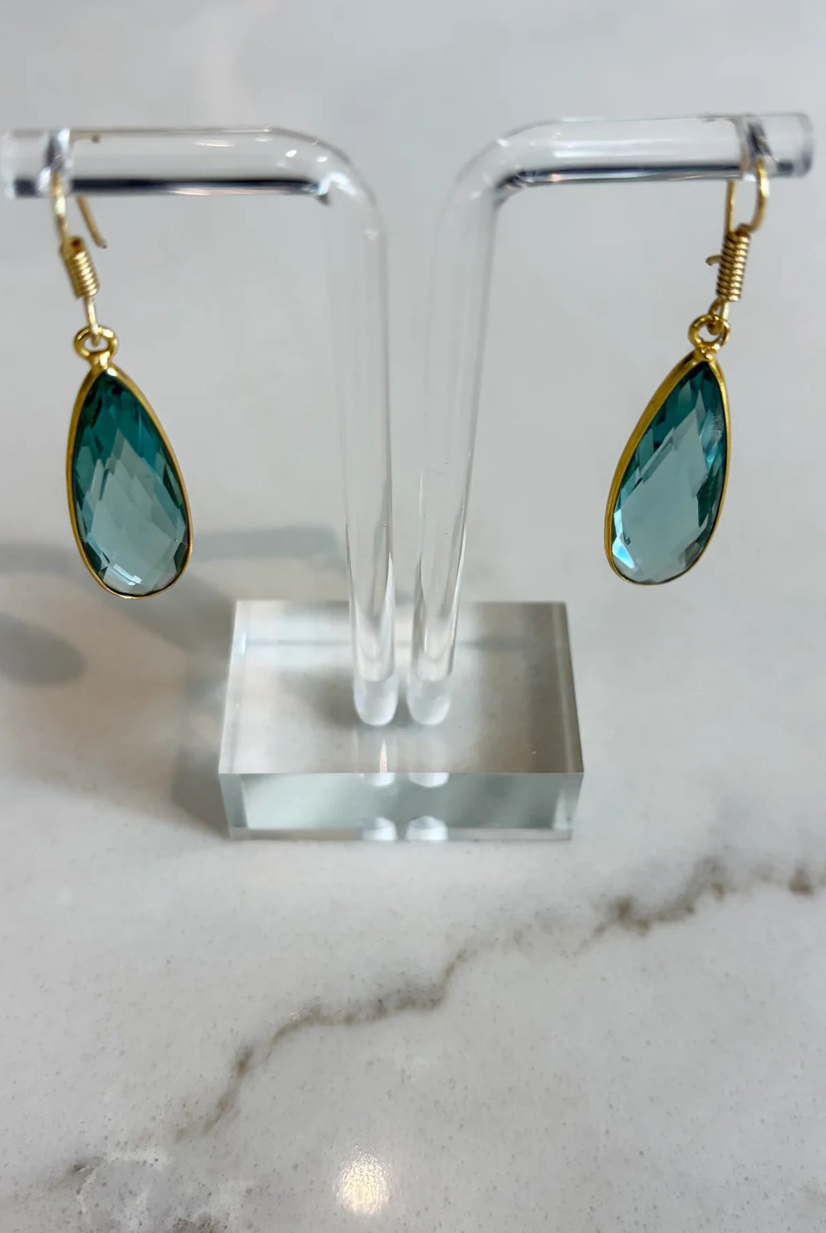 Teardrop-shaped earrings on a clear stand with a marble background. A pair of handcrafted soft teal Aurea drop earrings by Emilia Merz, showcasing the light-catching facets and gold hook closure.