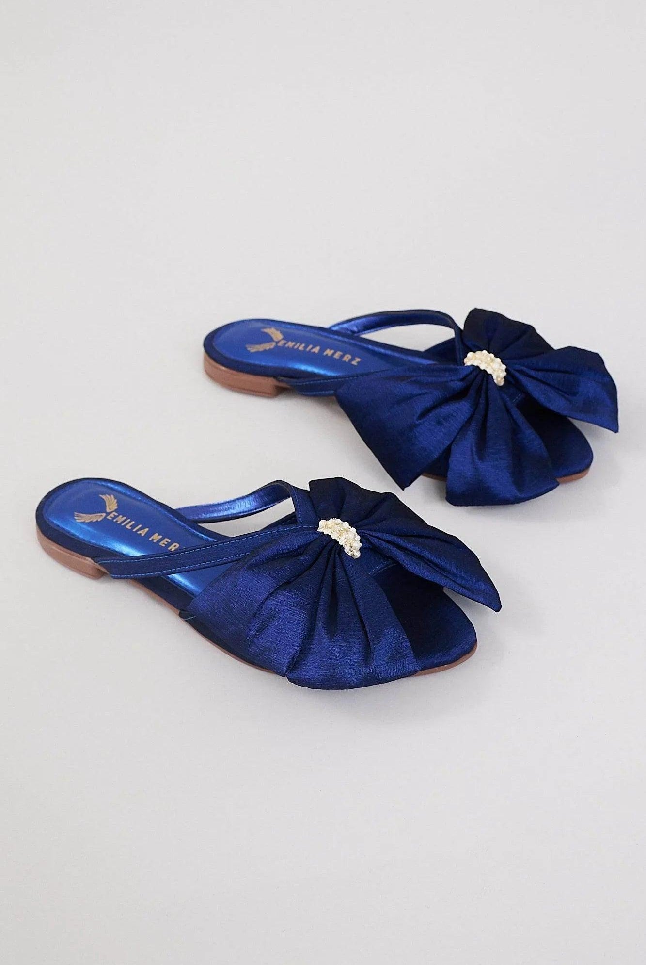 Navy blue flats with large satin bows and pearl embellishments on a neutral background.
