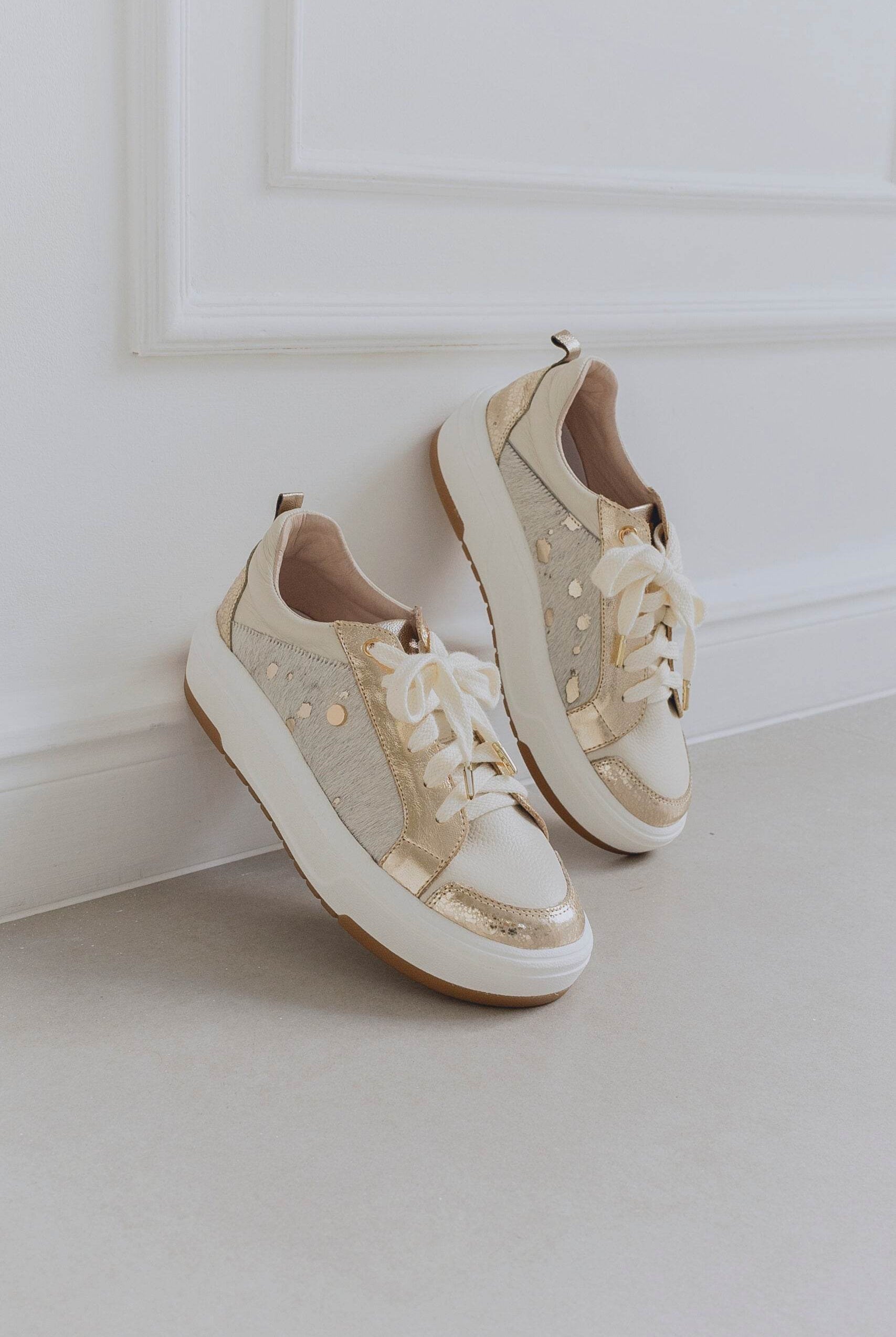 Pair of beige sneakers with gold accents on a light gray floor against a white wall.