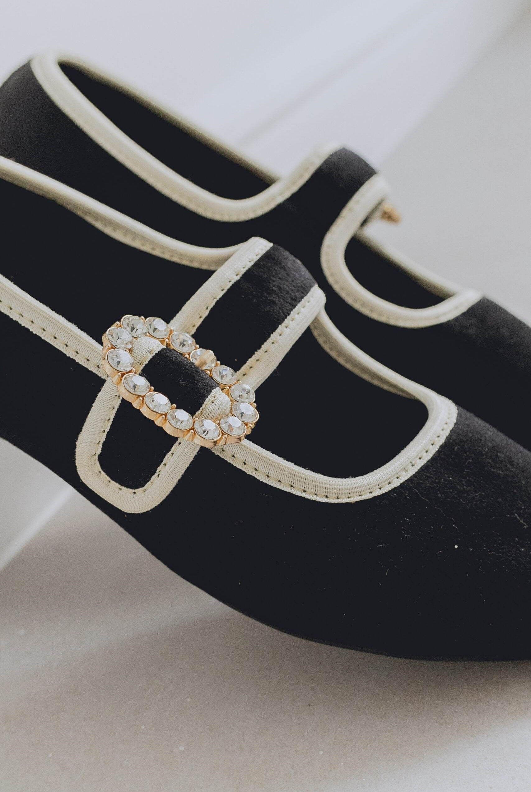 Black ballet flats with a decorative strap featuring a oval-shaped brooch on a light background