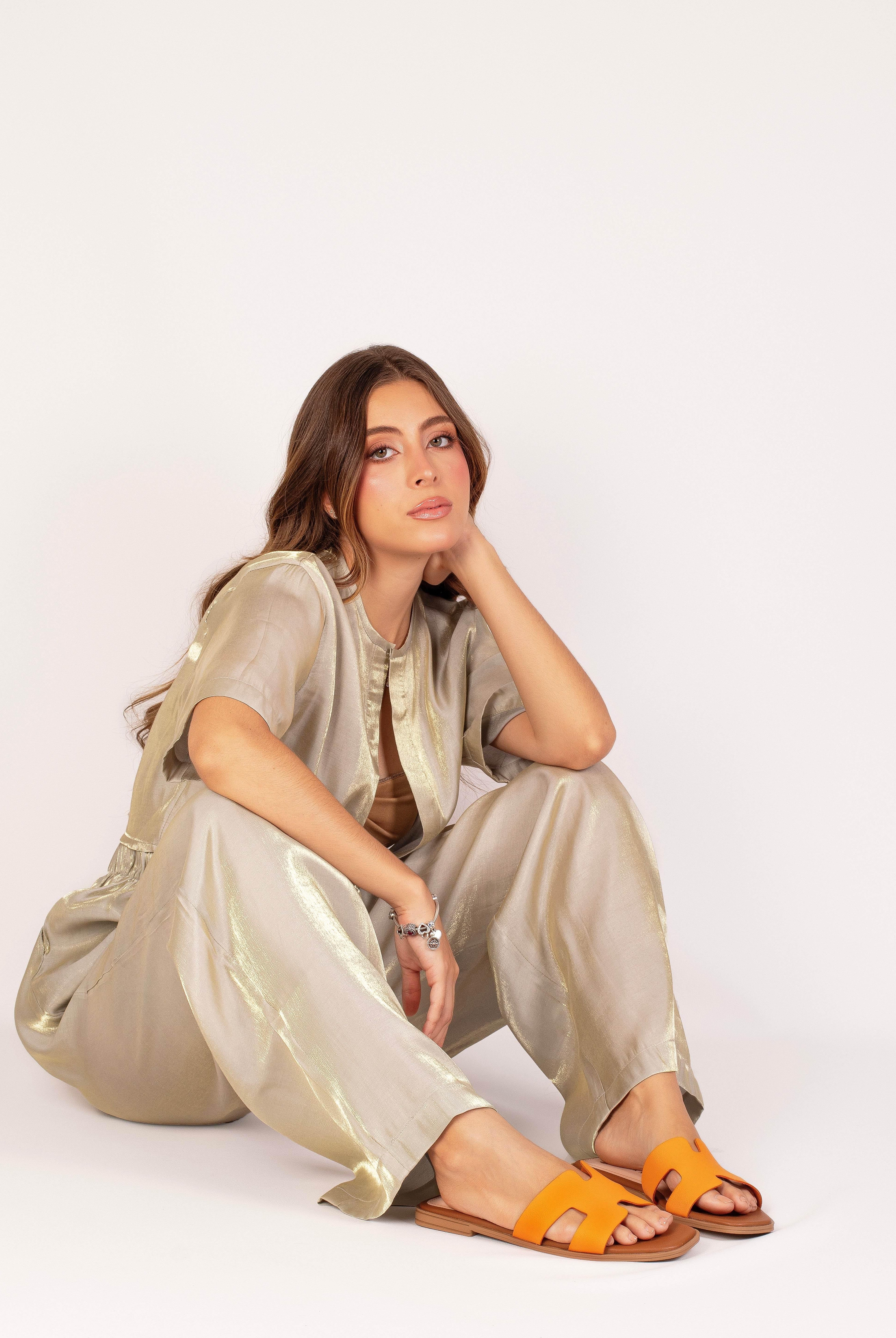 Woman in metallic outfit sitting, wearing orange H-strap sandals against white background