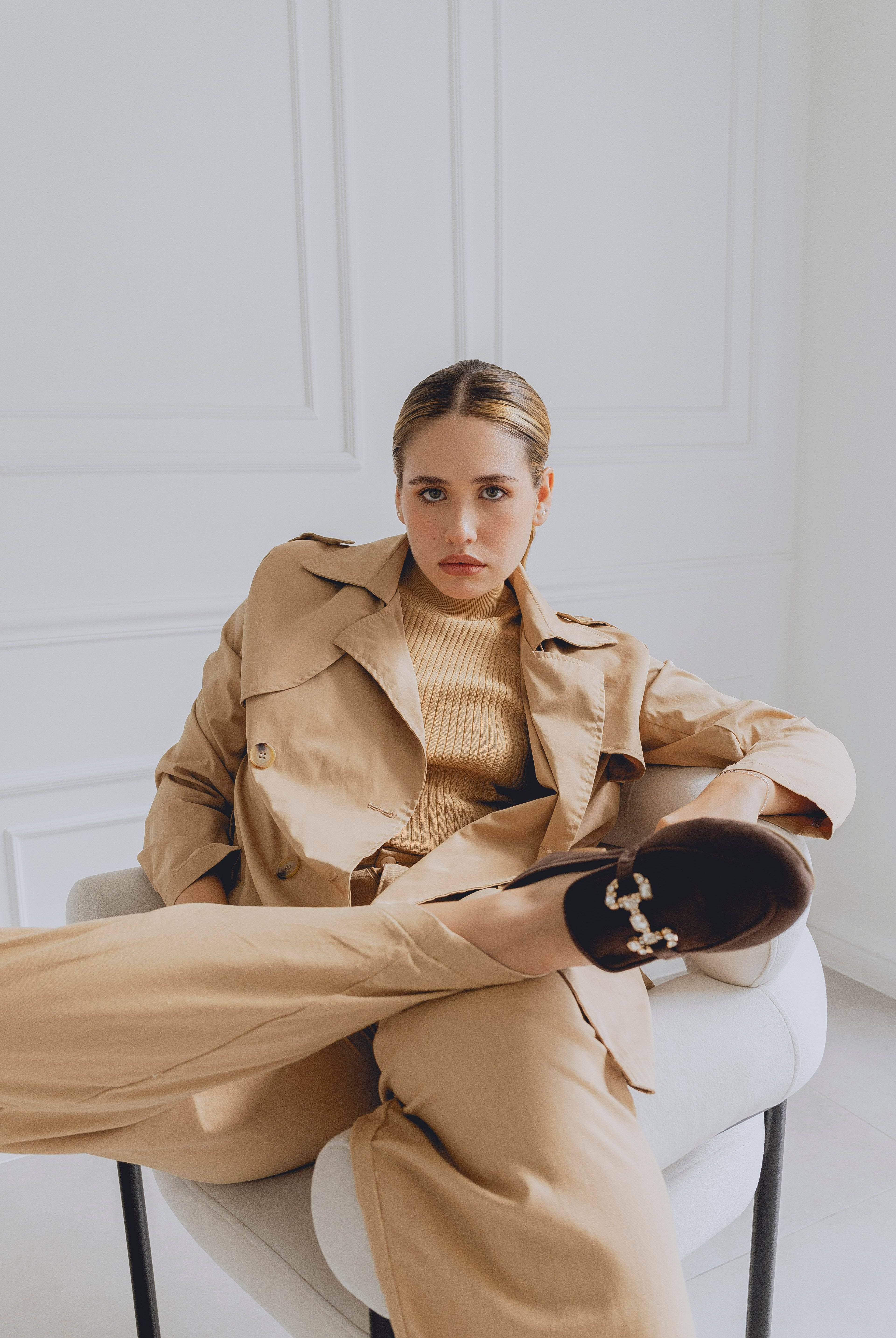 Woman in a beige trench coat and brown loafers sitting on a chair in a minimalistic room.