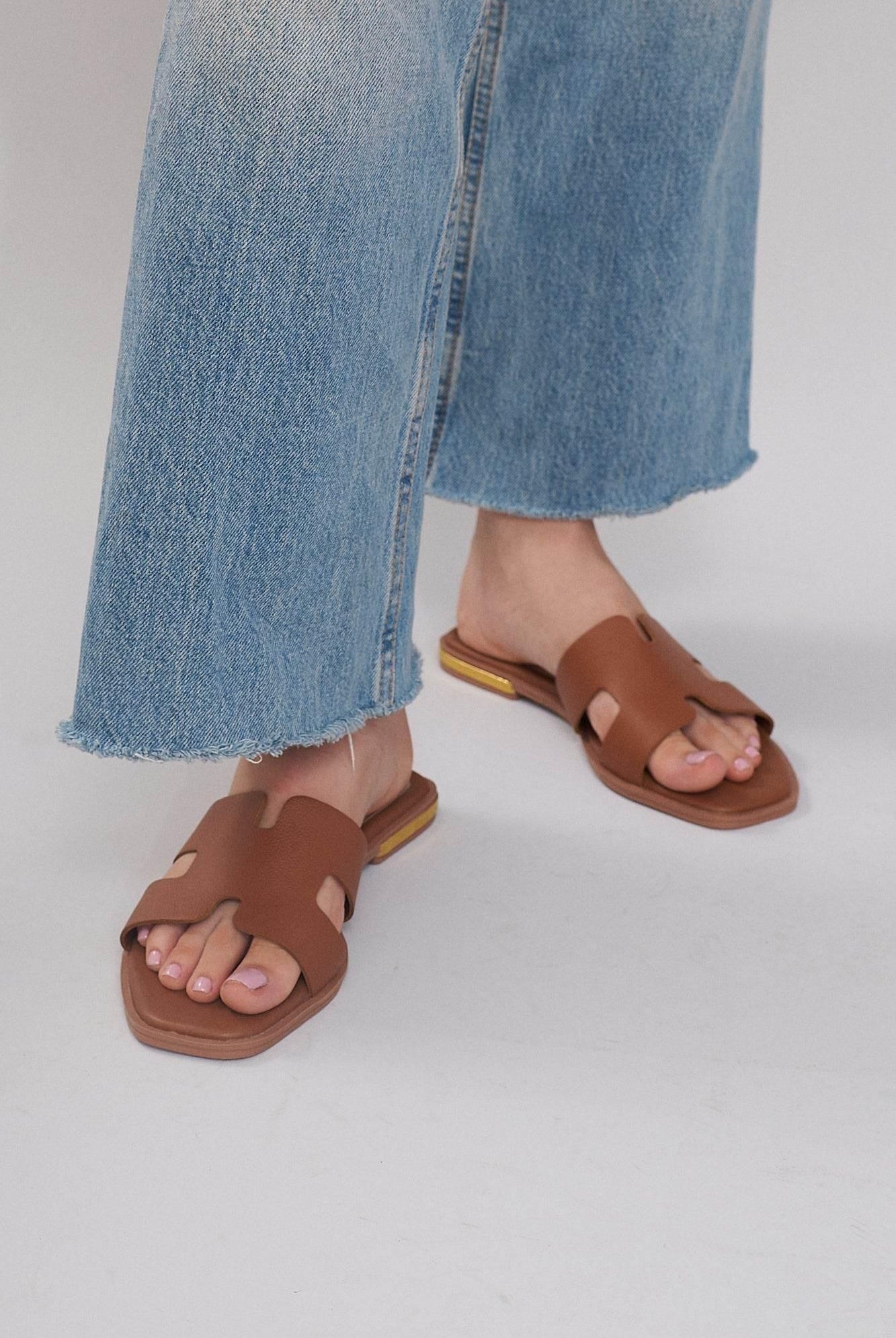 Brown leather slide sandals with cutout design paired with cropped blue jeans on light background