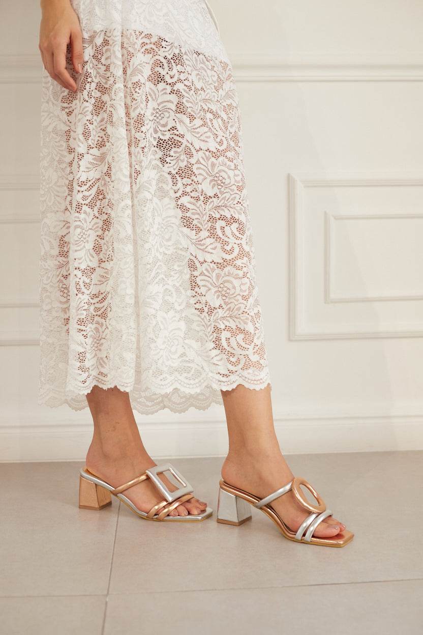 Person wearing white lace pants and rosegold and silver sandals on a light floor.