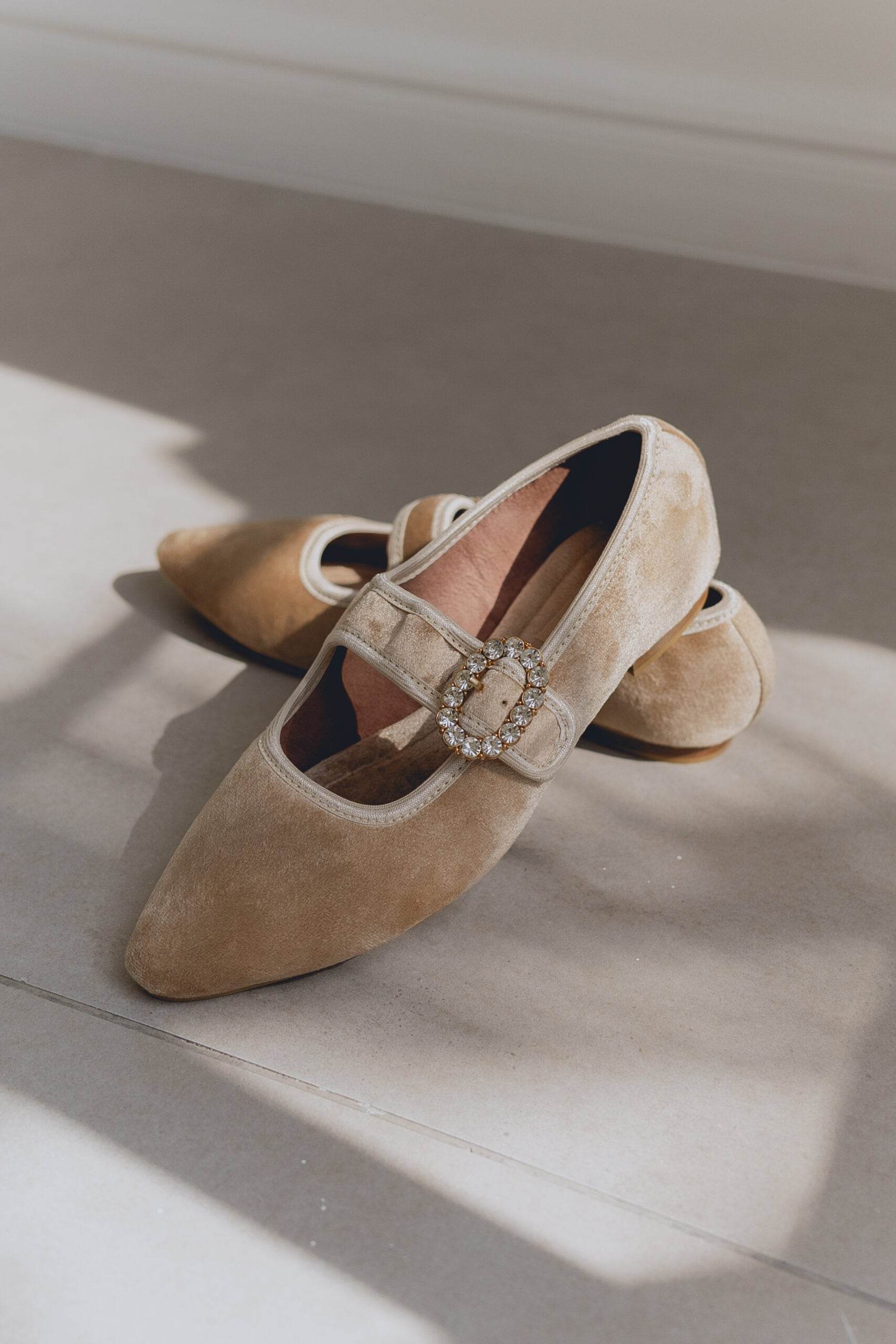 Pair of beige suede mary jane ballet flats with a decorative buckle on a light surface.