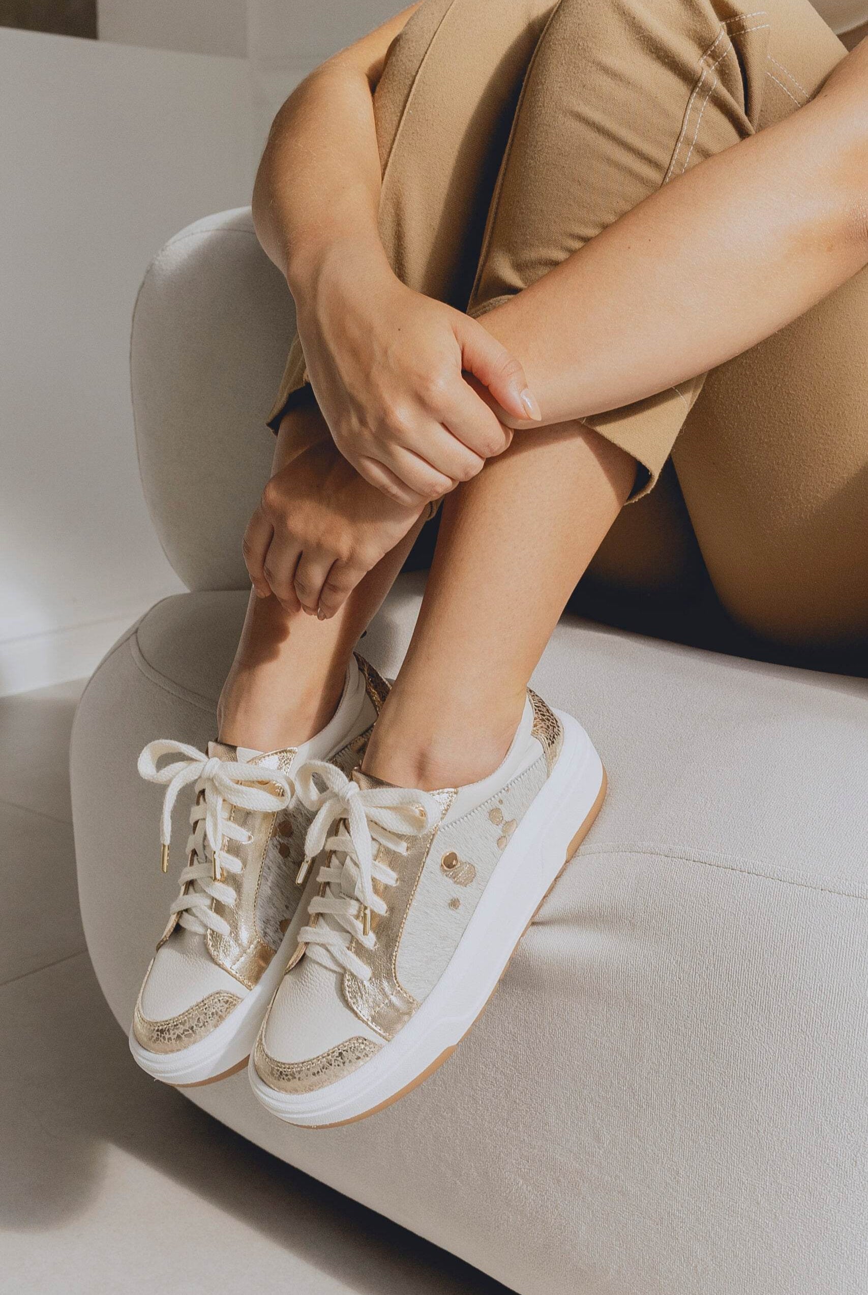 Person wearing beige and white sneakers with gold accents sitting on a light-colored couch.