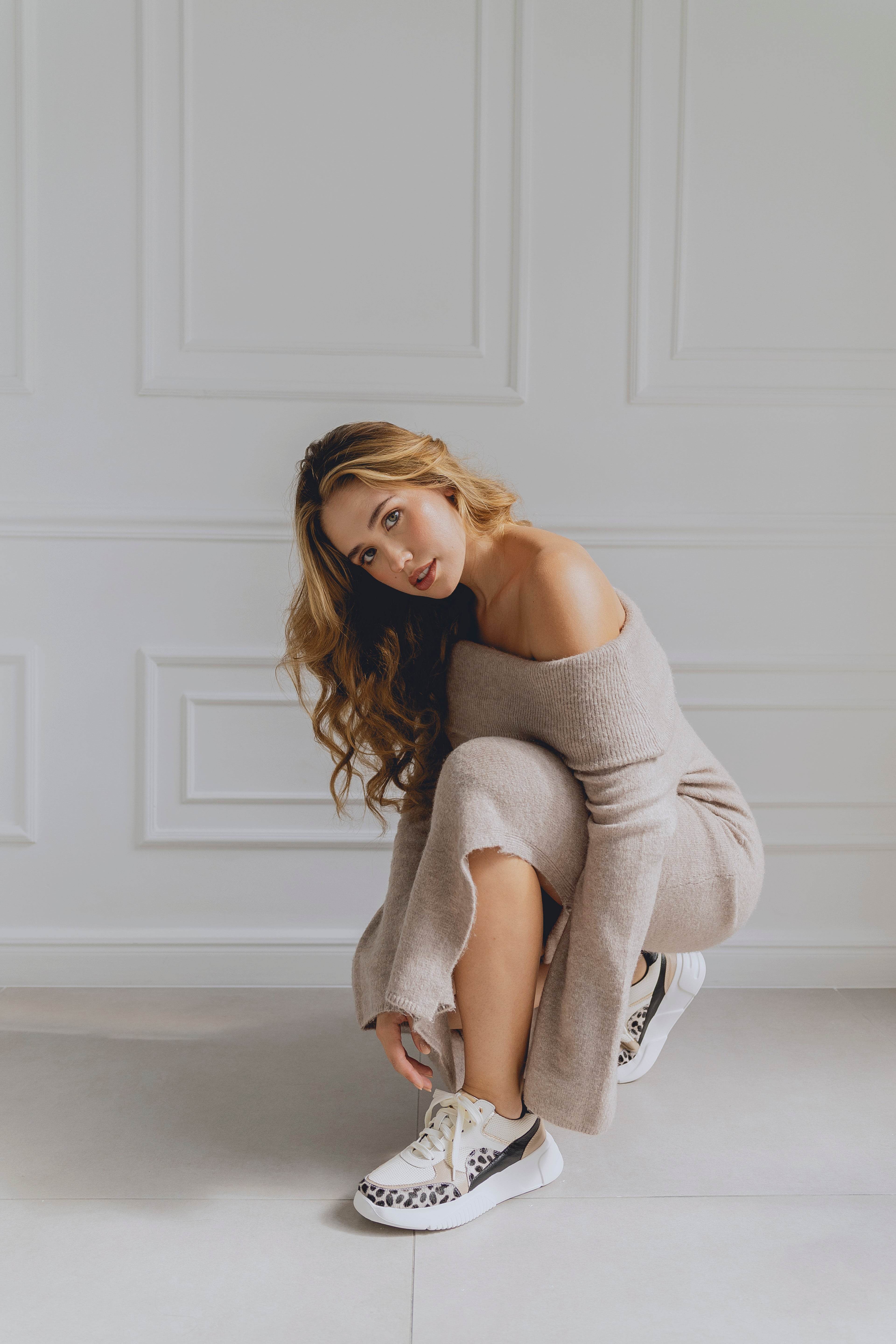 Woman in a beige off-shoulder dress with white sneakers sitting on a light-colored floor against a white paneled wall.