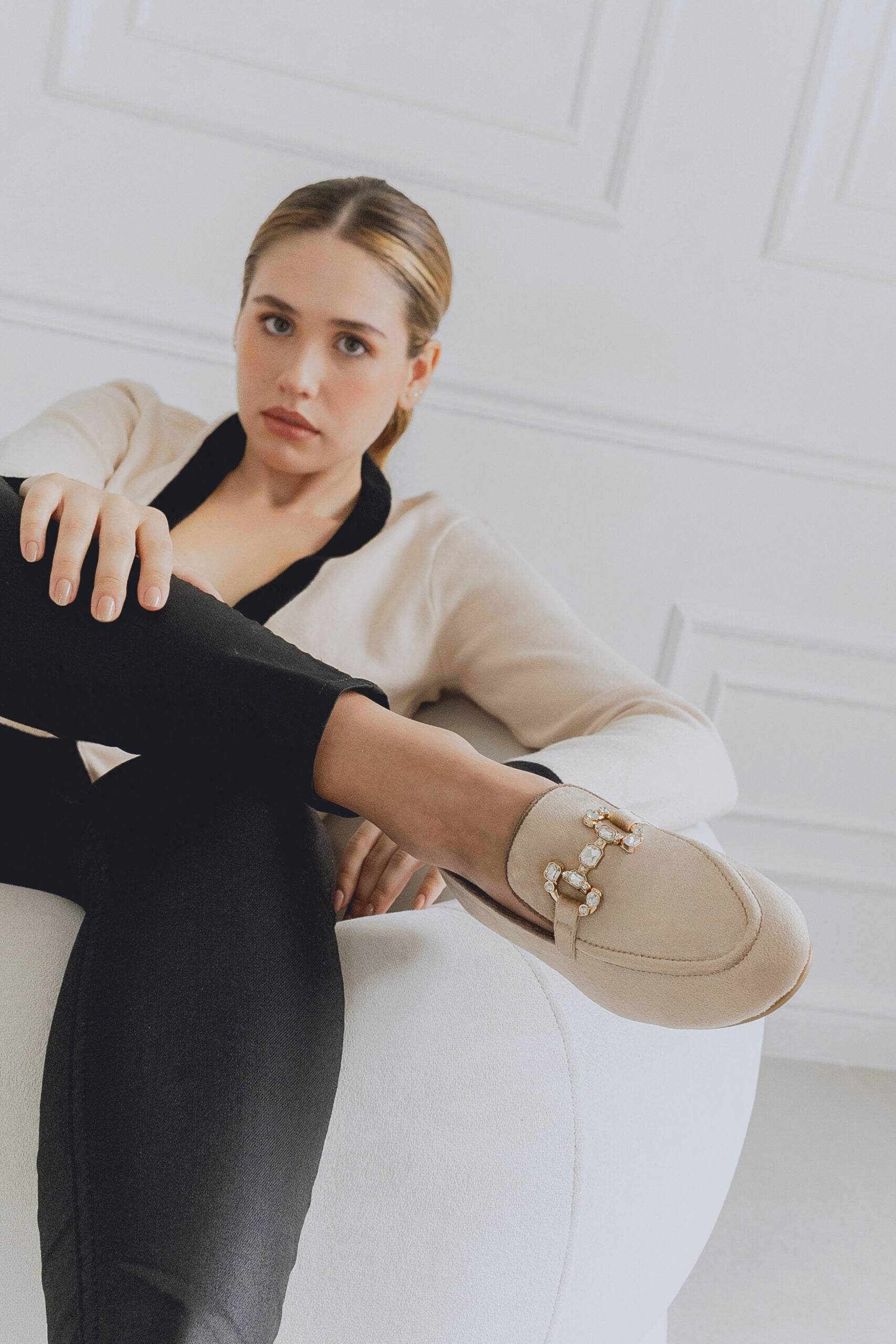 Woman wearing beige loafers sitting on a white surface with a white wall background