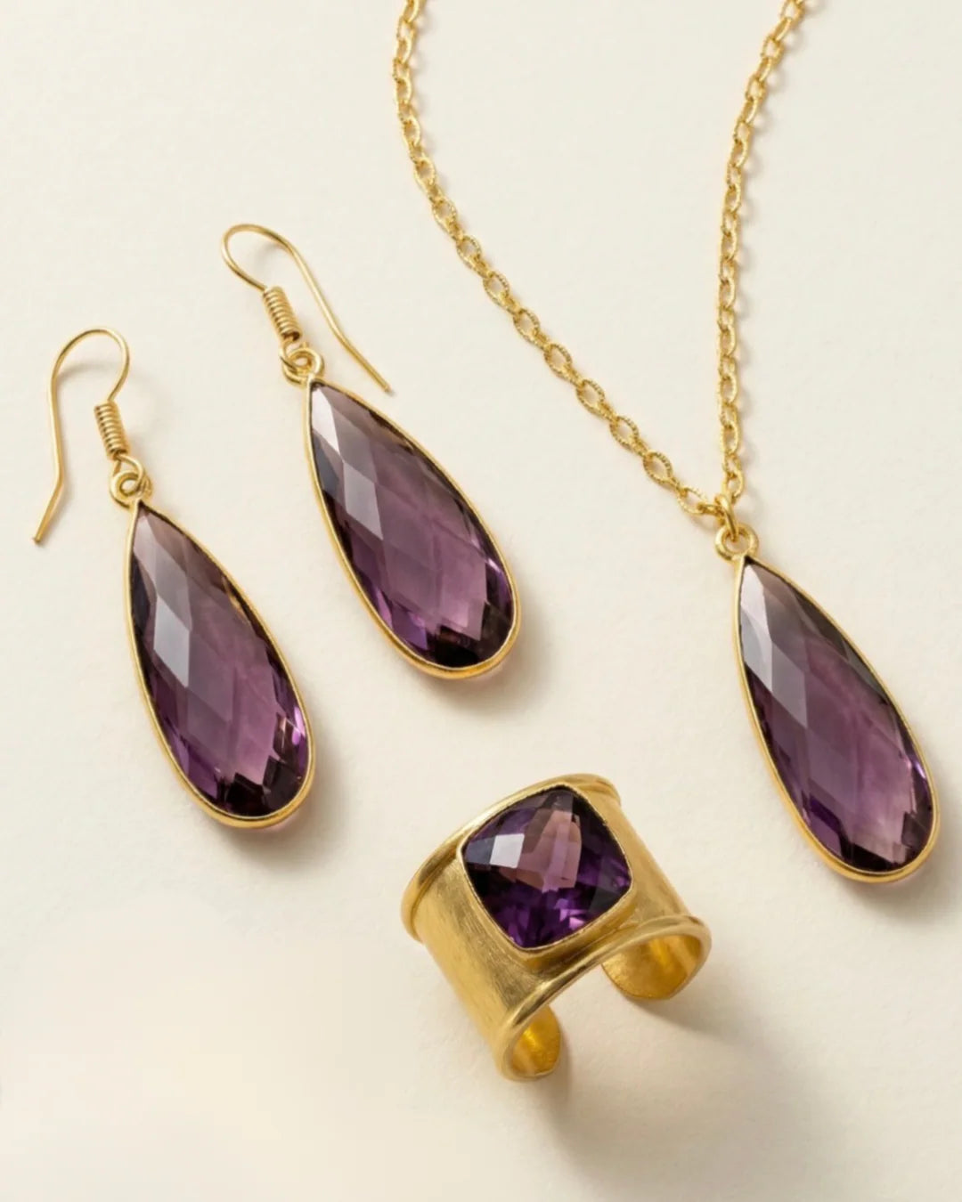 Emilia Merz Aurea jewelry set featuring a pair of faceted deep violet drop earrings, a matching pendant necklace, and a cuffed statement ring set in 18K gold plating on a light background.