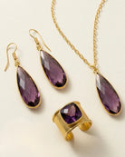 Emilia Merz Aurea jewelry set featuring a pair of faceted deep violet drop earrings, a matching pendant necklace, and a cuffed statement ring set in 18K gold plating on a light background.