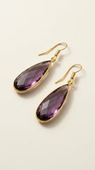 Gold aurea drop earrings with deep violet gemstones on a beige background
