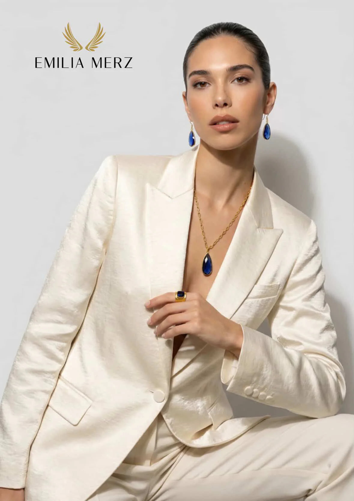 Woman in a white suit with emilia merz jewelry, wearing blue aurea earrings and a aurea necklace, on a light gray background.