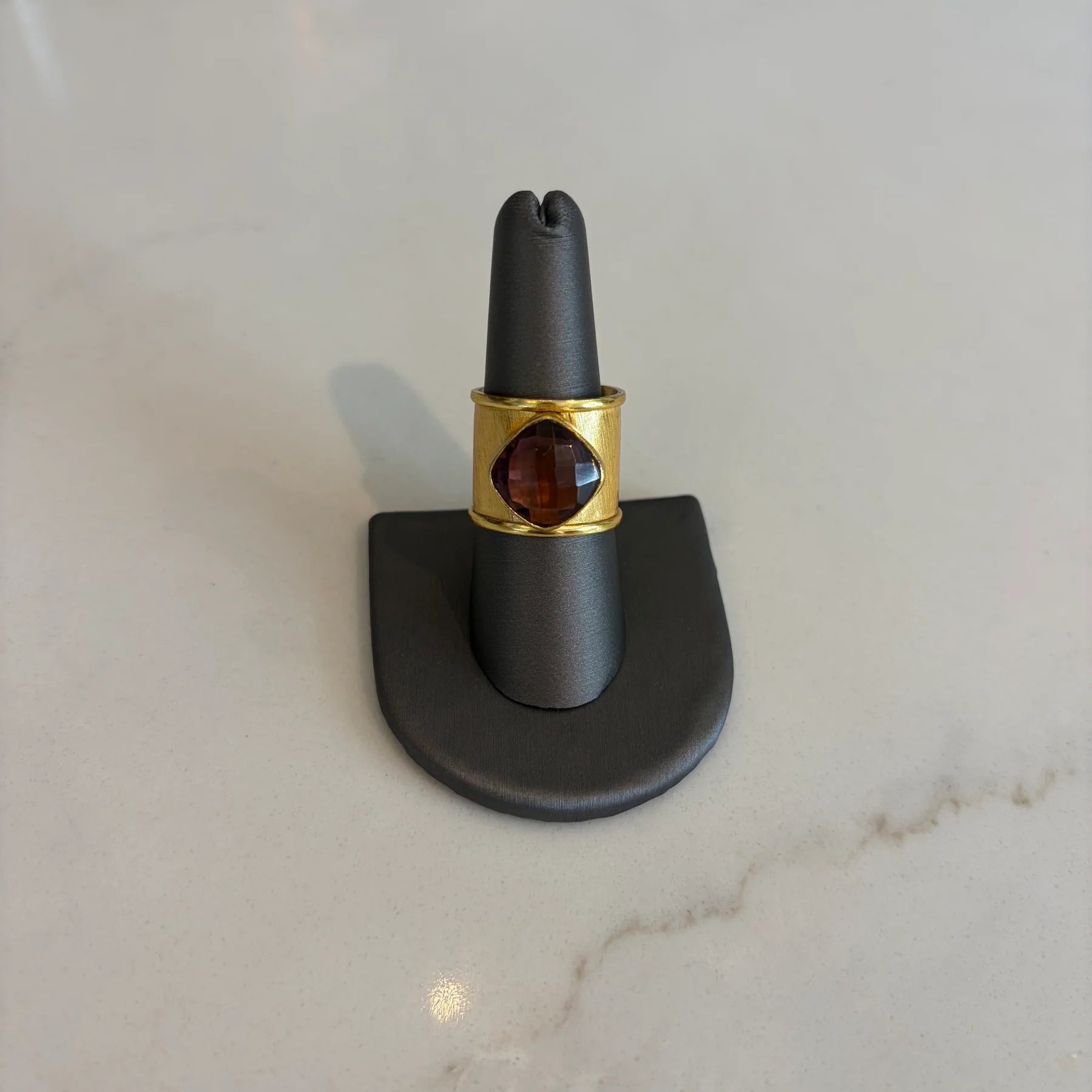 Front view of the Emilia Merz Aurea ring featuring a deep violet cushion-cut faceted stone set in a wide, cuffed 18K gold-plated band on a gray jewelry display.