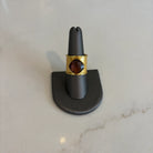 Front view of the Emilia Merz Aurea ring featuring a deep violet cushion-cut faceted stone set in a wide, cuffed 18K gold-plated band on a gray jewelry display.