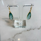 Teardrop-shaped earrings on a clear stand with a marble background. A pair of handcrafted soft teal Aurea drop earrings by Emilia Merz, showcasing the light-catching facets and gold hook closure.