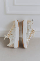 Pair of white sneakers with brown soles and gold spots on a light gray surface.