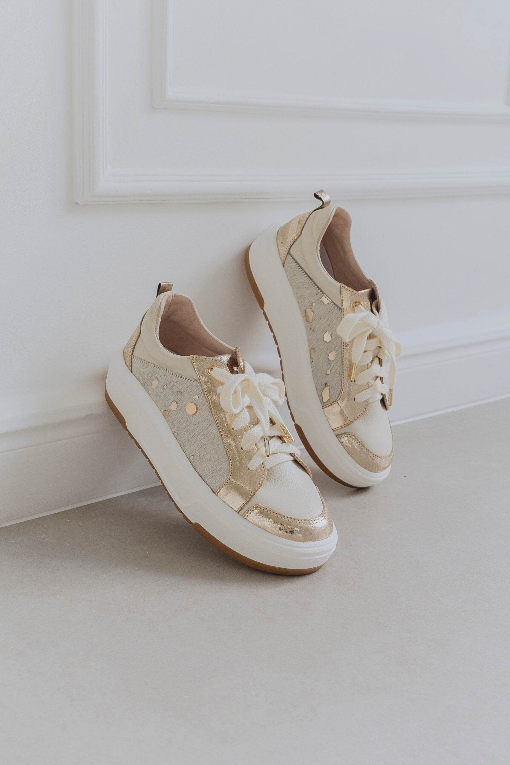 Pair of beige sneakers with gold accents on a light gray floor against a white wall.