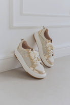 Pair of beige sneakers with gold accents on a light gray floor against a white wall.