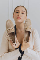 Woman holding two beige loafers with decorative buckles against a white background