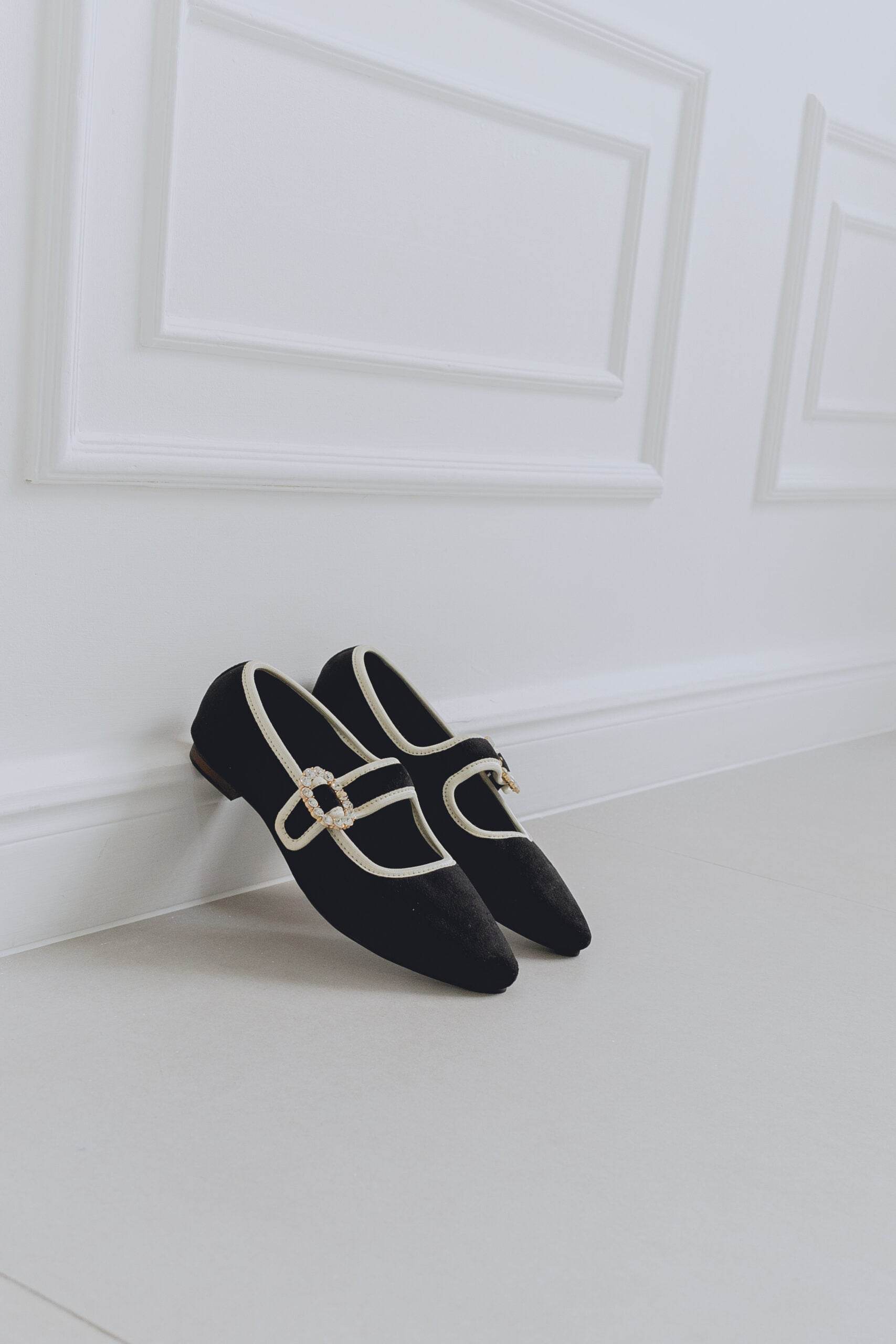 Black mary jane ballet flats with a gold buckle on a white floor against a white wall.