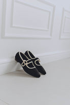 Black mary jane ballet flats with a gold buckle on a white floor against a white wall.