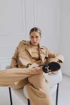 Woman in a beige trench coat and brown loafers sitting on a chair in a minimalistic room.