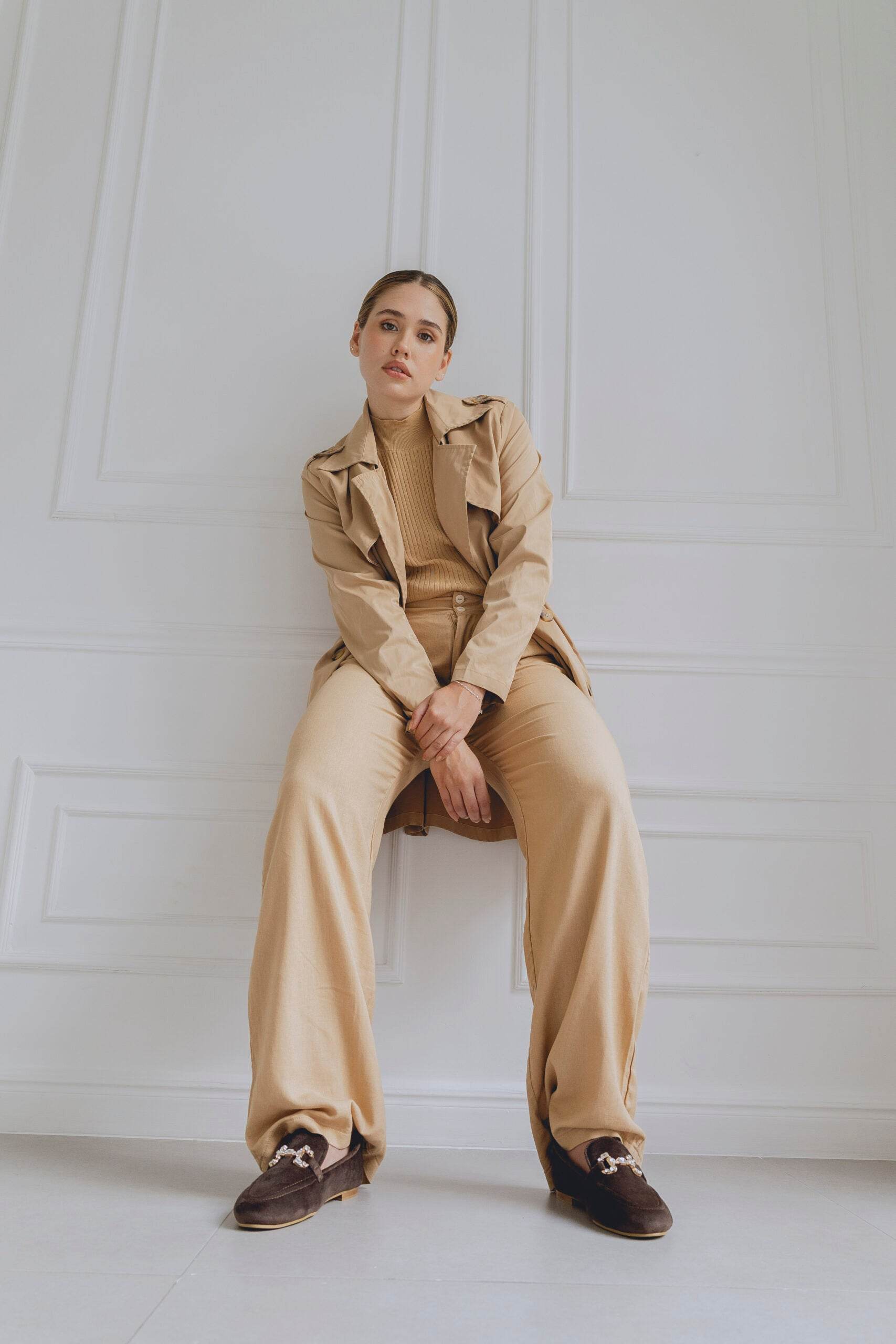 Person wearing a beige trench coat and pants with brown loafers sitting on a chair against a white wall.