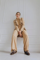 Person wearing a beige trench coat and pants with brown loafers sitting on a chair against a white wall.