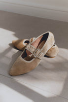 Pair of beige suede mary jane ballet flats with a decorative buckle on a light surface.
