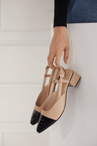 Beige slingback heels with black pointed toes held by a hand against a light background