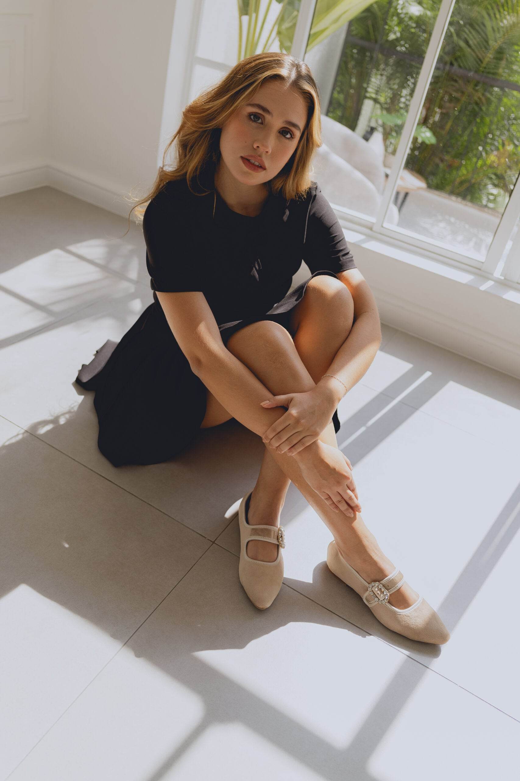 Woman sitting on a tiled floor in a bright room with large windows wearing beige ballet flats.