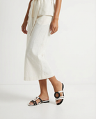 Person wearing a white outfit with asymmetrical sandals on a plain background