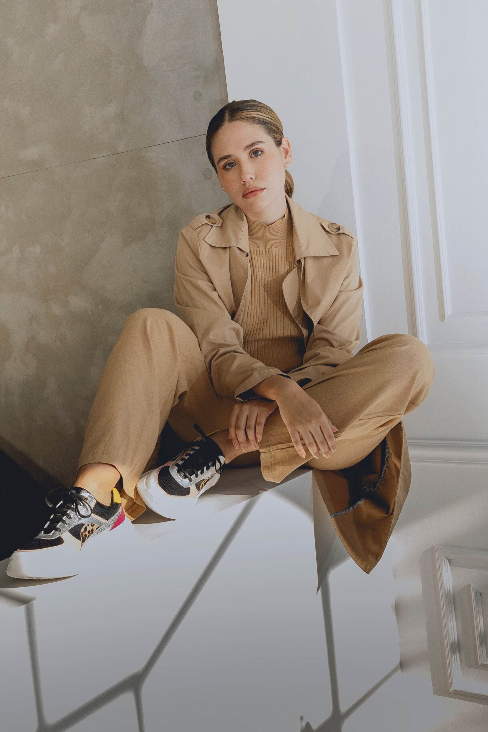 Woman in a beige trench coat and pants sitting wearing multi colored sneakers on a white floor.
