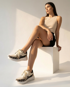 Woman sitting on a white block wearing beige sneakers with leopard print details.