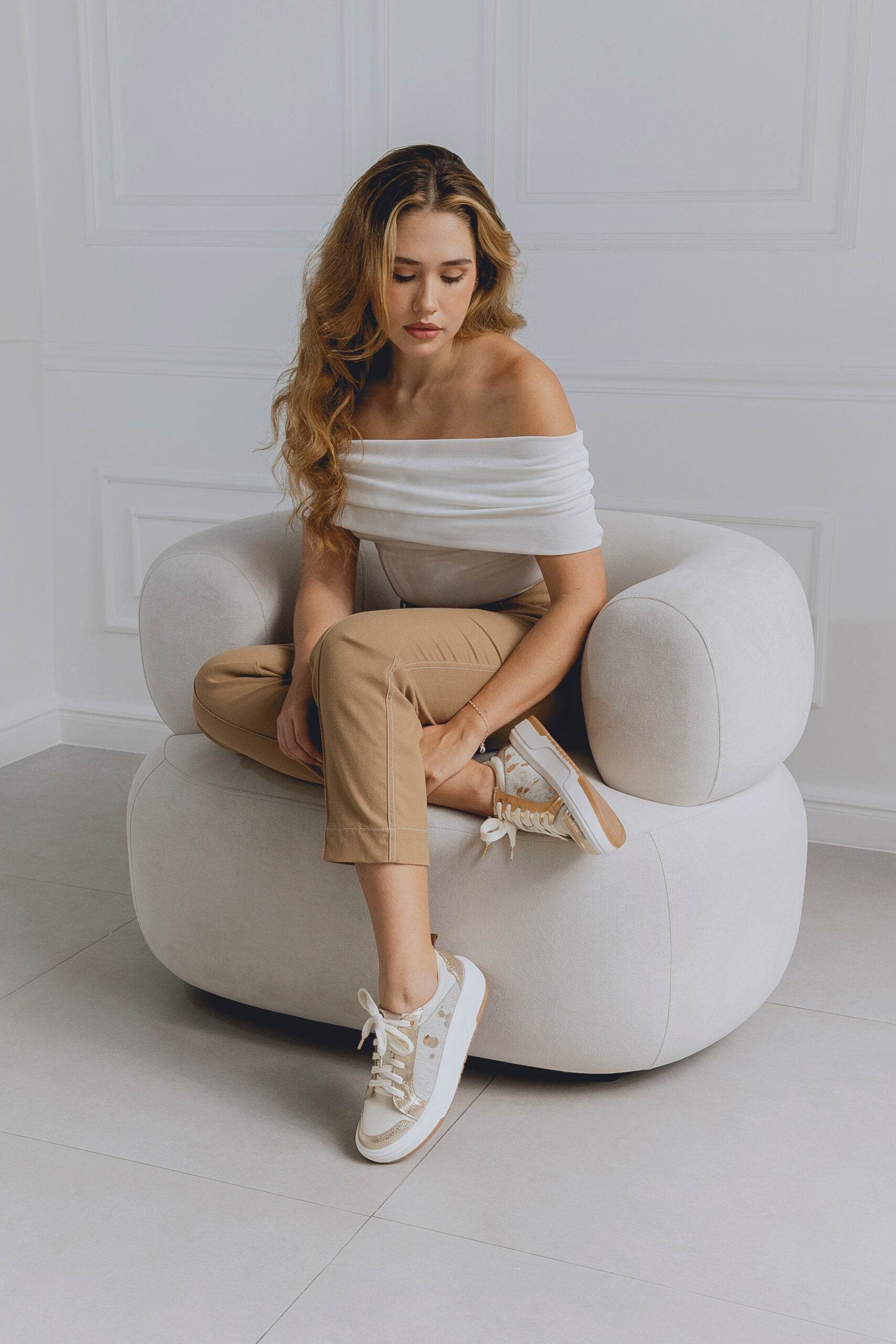 Woman sitting on a white chair in a minimalistic room wearing beige white sneakers for women with golden sprinkles