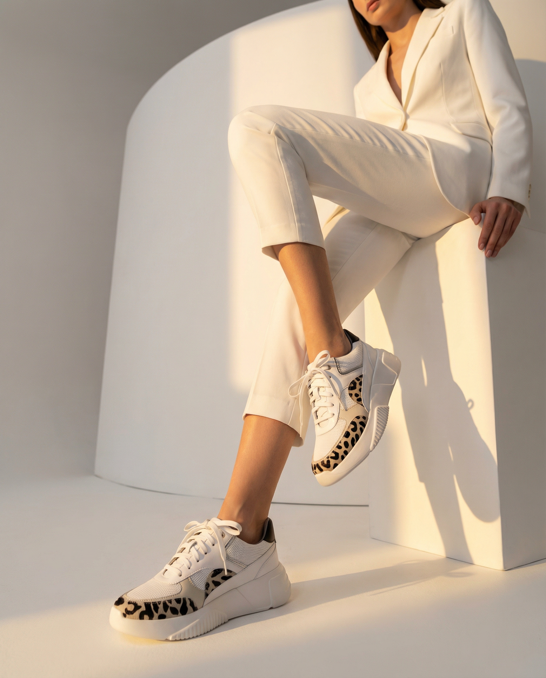 Person wearing white sneakers with black leopard print details, sitting on a white architectural structure.