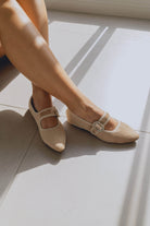 Beige ballet flats with decorative straps worn by a person sitting on a light-colored floor.