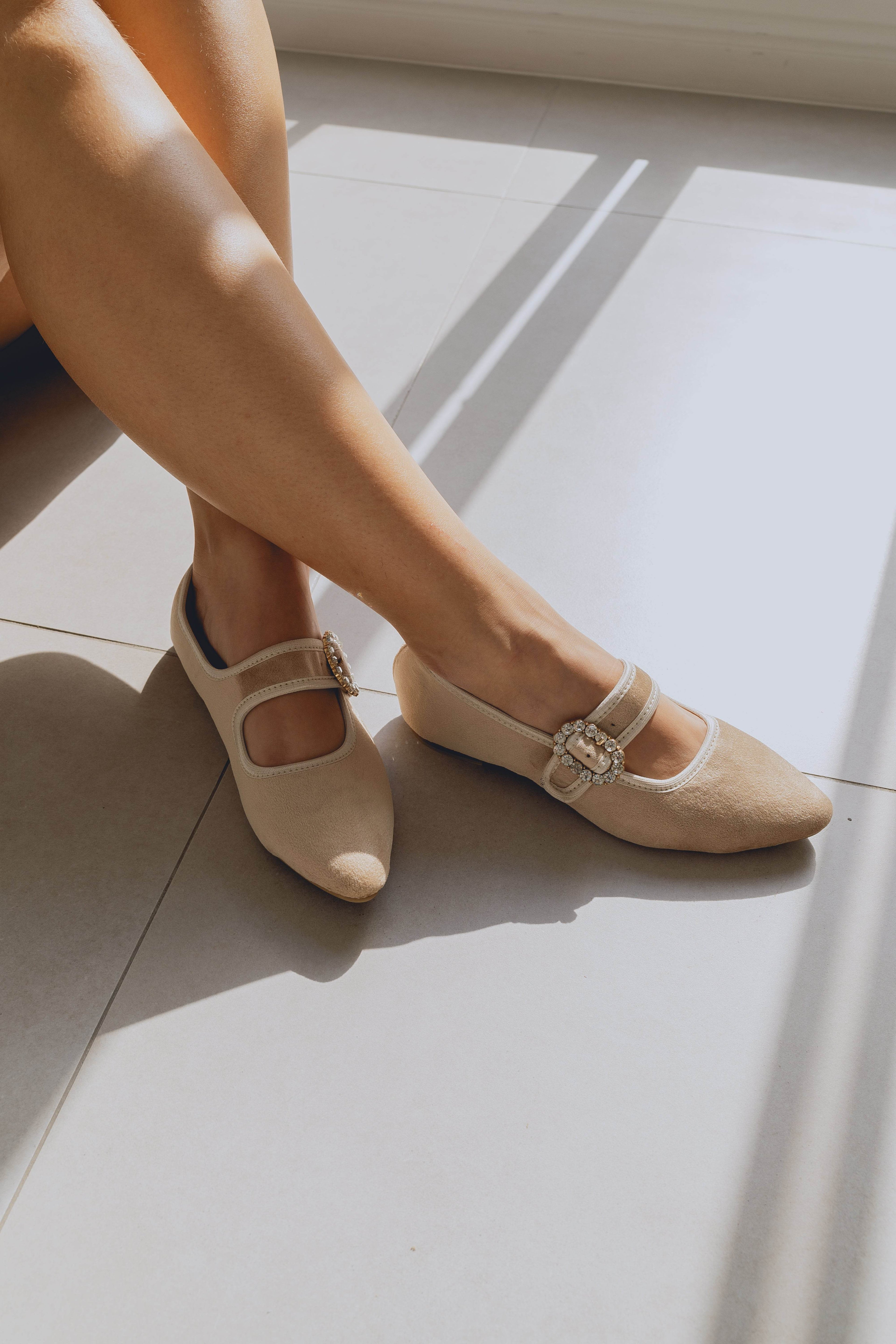 Beige ballet flats with decorative straps worn by a person sitting on a light-colored floor.